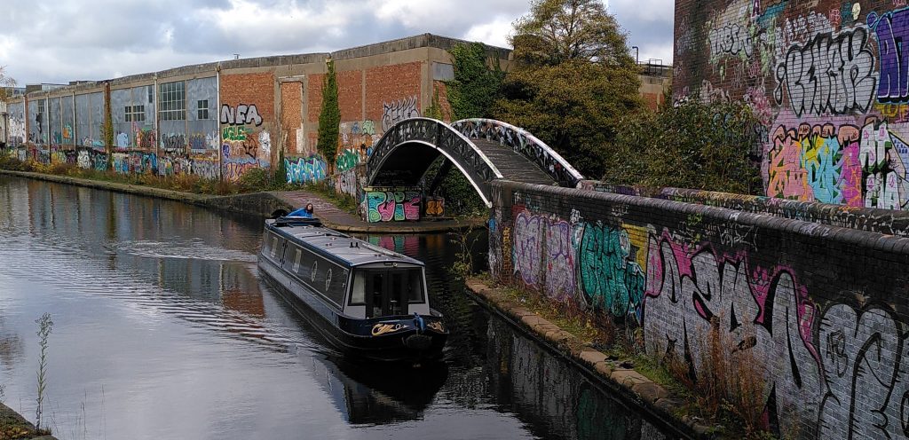 Clare driving Bartimaeus past a black and white painted junction bridge surrounded by brick factories covered in graffiti.