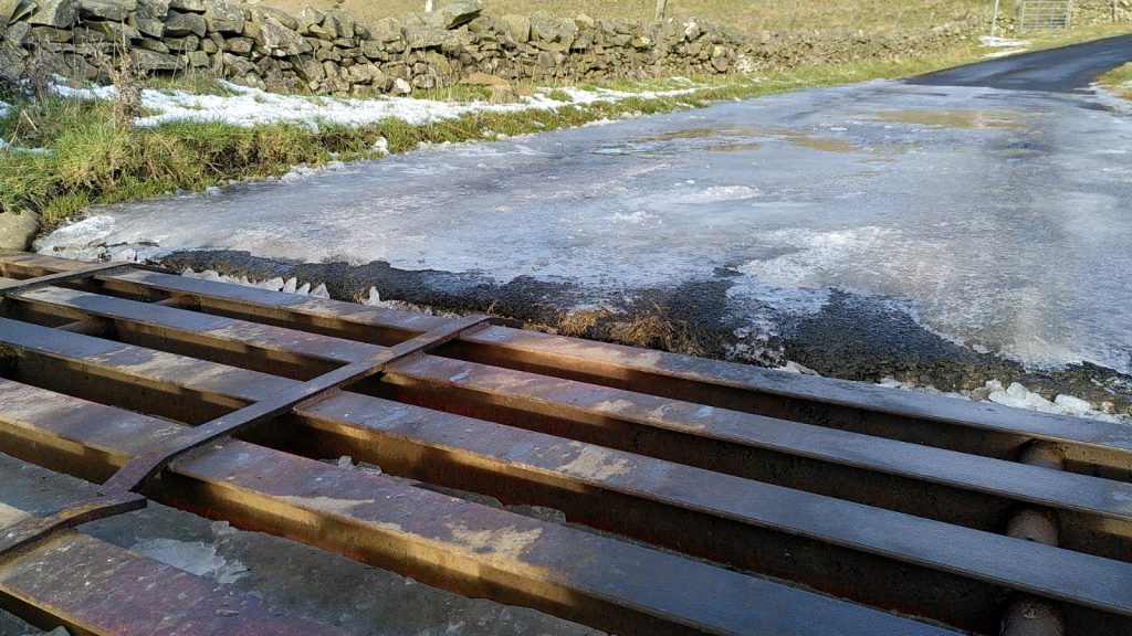 Cattle grid with ice under the bars and a sheet of ice over the road beyond.