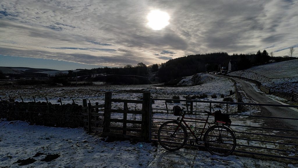 View of weak sun shining over snow covered hills. A bicycle is leaning on a gate in the foreground.