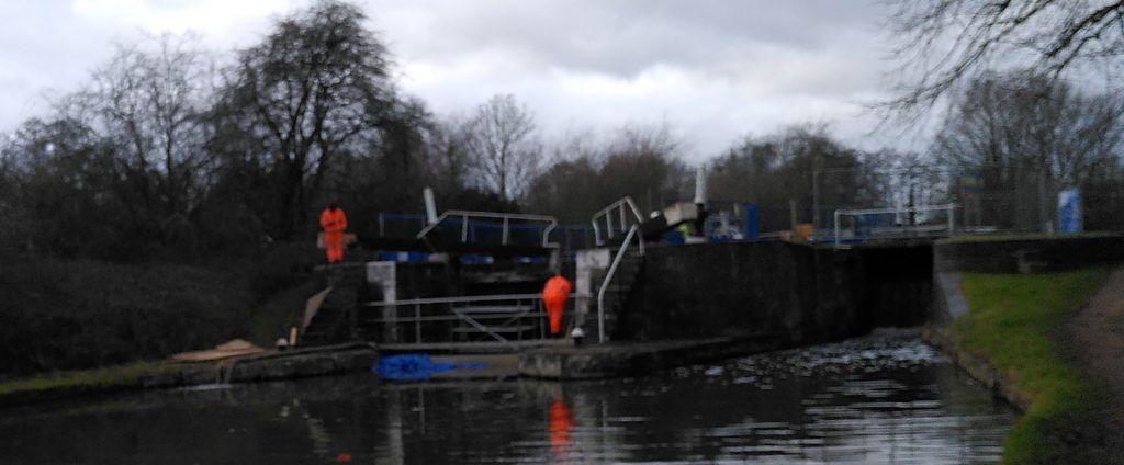 Two workers in orange on either side of open lock gates seen across a canal.