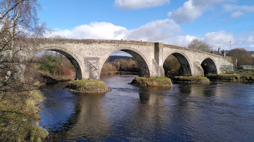 Multi arch stone bridge over the River Forth.  Blue sky is reflected in the river. The Wallace Monument is just visible above the far end of the bridge.