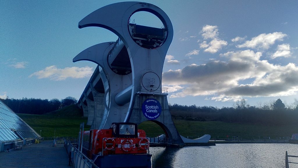 Trip boat moored beside the Falkirk Wheel.  The wheel towers above, its ends looking like enormous beaked heads.