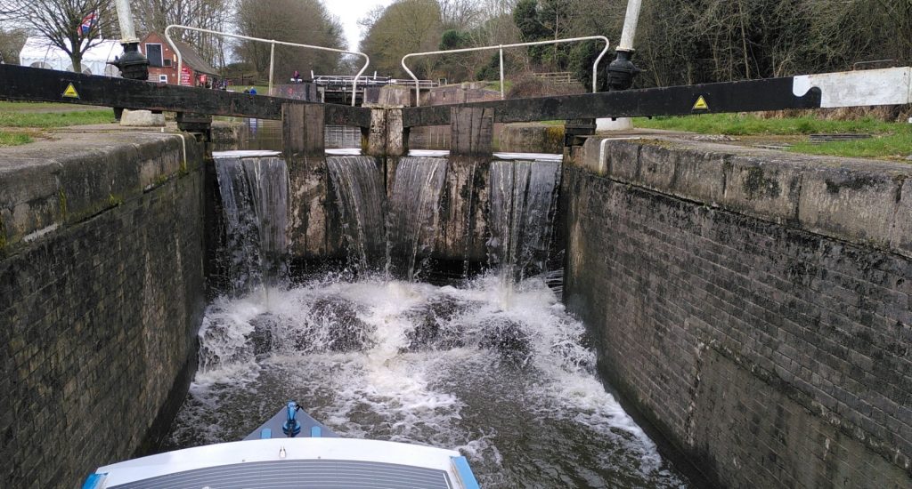 Water cascades over the head gates of a double lock.  In the foreground is the bow and roof of a narrowboat.