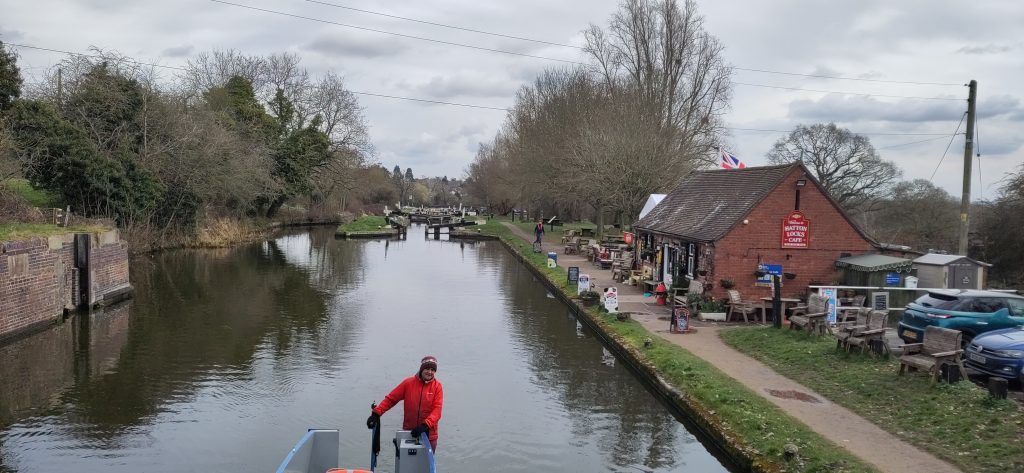 A wide canal with a cafe on the towpath side.  In the distance, a lock gate is visible, in the foreground a narrowboater steers under the viewer.