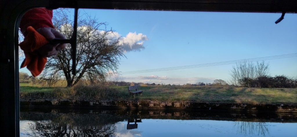 Canal and blue sky beyond viewed through a window.  A hand is wielding a cleaning wiper in one corner of the window.