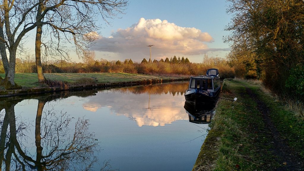 A still canal reflects a blue sky in which hangs a pink and white cloud.  A narrowboat is moored against the towpath in the foreground.