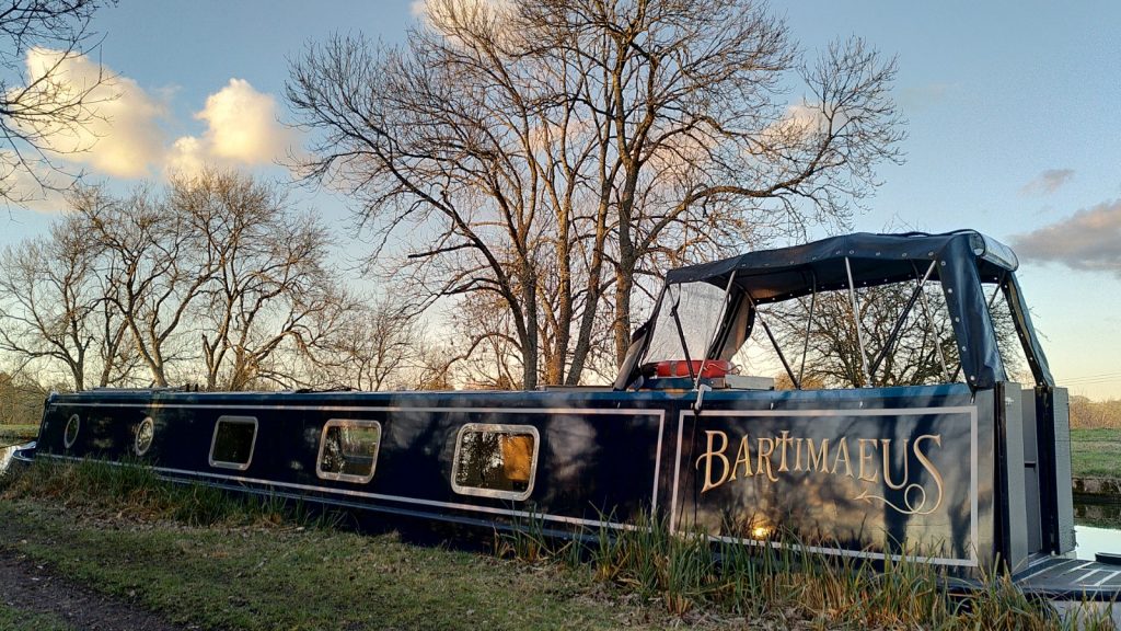 A moored narrowboat seen side on with a golden hue added by a low sun. Bare trees behind partially obscure a blue sky with fluffy clouds.