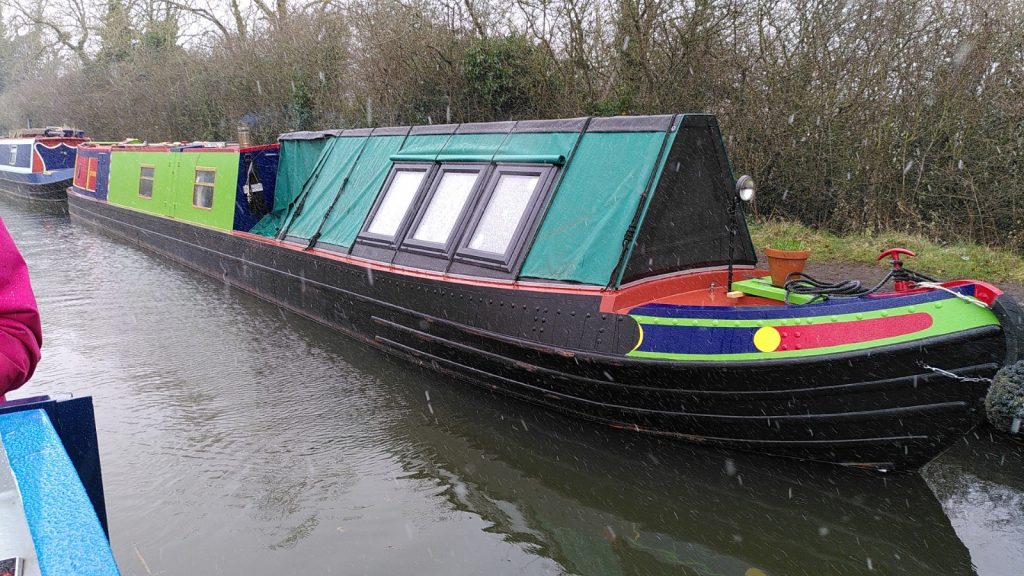 Moored narrowboat in bright colours.  The front section of the boat is a traditional A-frame covered in canvas, except that there are modern windows inserted in to the canvas.