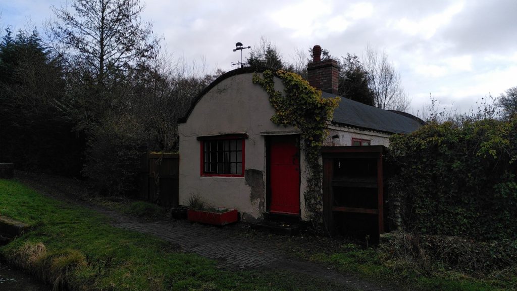 Lock keeper's cottage painted white.  The gable end faces the canal with a single window and a red door.  The roof is a characteristic barrel shaped.