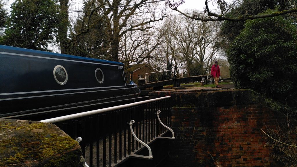 A narrowboat sits in a cast iron aqueduct.  A figure is operating the lock gate at one end of the aqueduct.  Details in the black painted aqueduct are picked out in white paint.