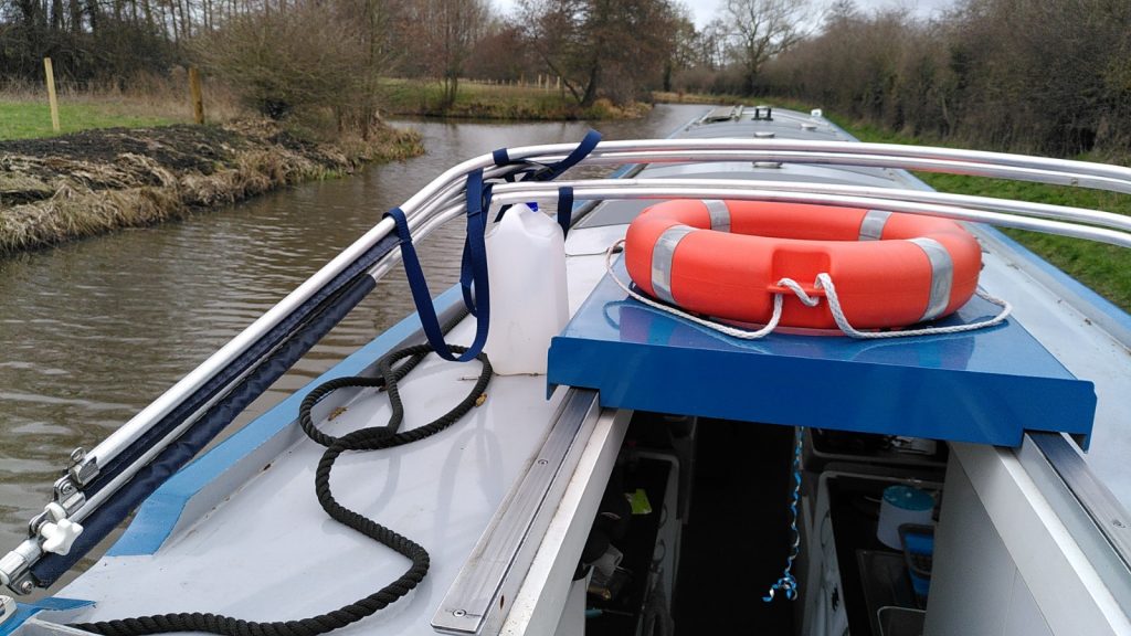 View from stern deck of narrowboat.  The hatch, with a life ring on top, is slid forward under some poles. The poles are propped p by a plastic bottle.