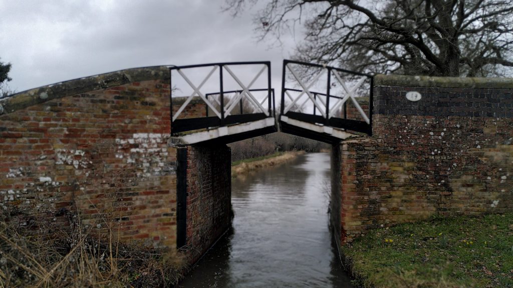 A typical South Stratford Canal bridge.  A square brick support sits either side of the canal. On top of each, a cast iron plate angled up at about 15°. The plates do not quite meet.  Each has a handrail and is picked out in black and white paint.
