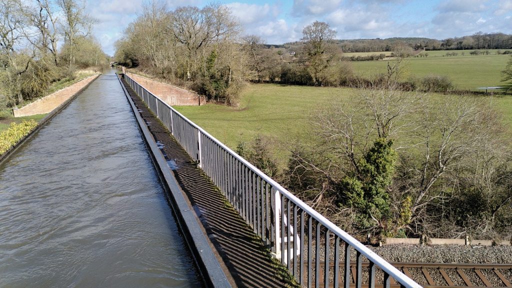 A cast iron trough takes the canal to a vanishing point.  To the right the towpath is at the level of the bottom of the canal with a white painted fence. There is no fence for the boats. A railway line is just visible on the ground below.