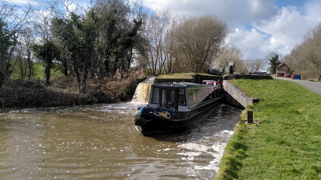 A narrowboat is exiting the lower gate of a lock. On the far side of the boat a waterfall cascades in to the brown canal water with an apparent height similar to the boat.