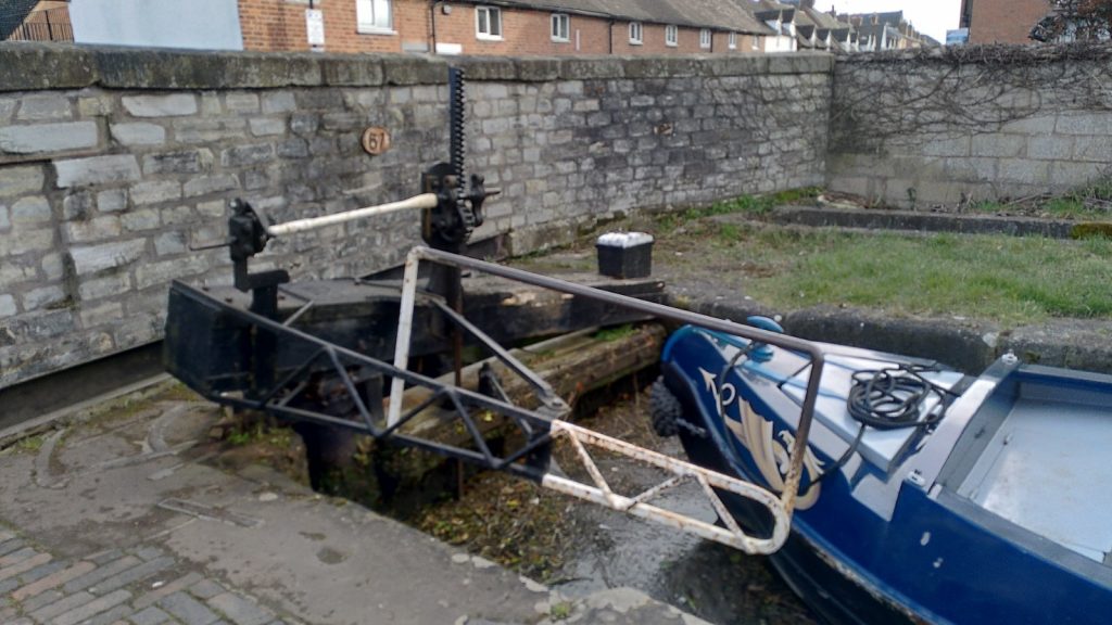 The nose of a narrowboat sits against a lock gate.  The arm of the lock gate has been replaced by a metal frame at right angles to the gate.  It is not as long as the canal is wide. Beyond the lock gate is a stone wall on a bridge over the canal.