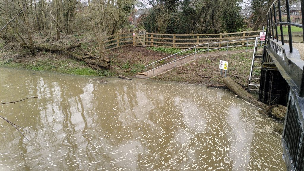 On the far side of a stretch of water, a sloped path leads into the water's edge. The only sign of the submerged walkway leading along the shore is a barely-visible post top. A large tree trunk is lying across the lock entrance against the gates.