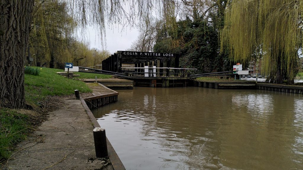 Willow branches droop over the river above a lock landing in the foreground. Small posts for tying boats are clearly visible.  The river stops at double gates to a lock . A metal gantry over the lock bears the words "COLIN P. WITTER LOCK".