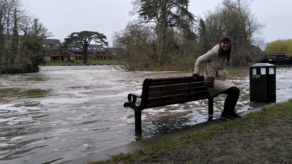 A women leans against the back of a bench.  The bench faces the river, but its feet are in the flood.  Across the river a wide weir flows straigh toards the bench.