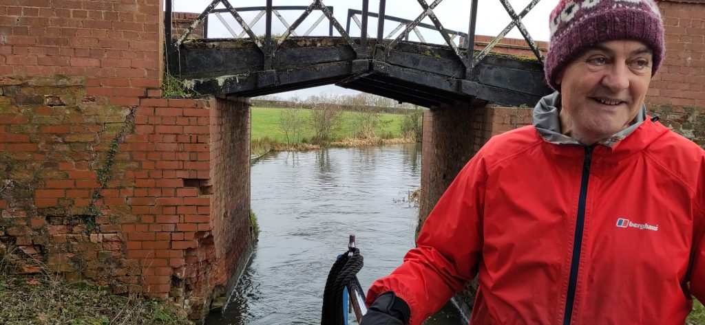 Shane driving Bartimaeus in hat and gloves. Behind is a narrow bridge with brick parapets. One parapet has had many bricks knocked out near the waterline.