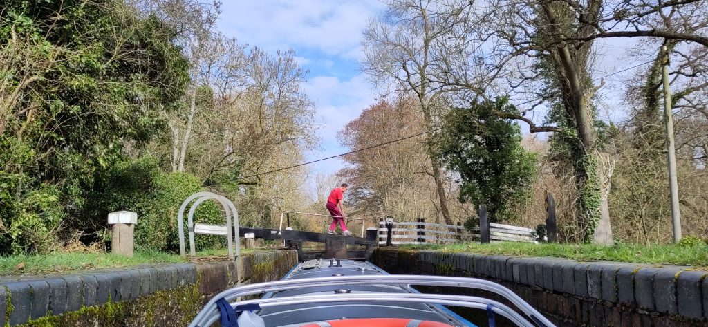 A figure in red crosses a lock gate seen along the roof from the other end of a narrowboat. The sky is blue with white clouds. The trees on either side are mostly bare.