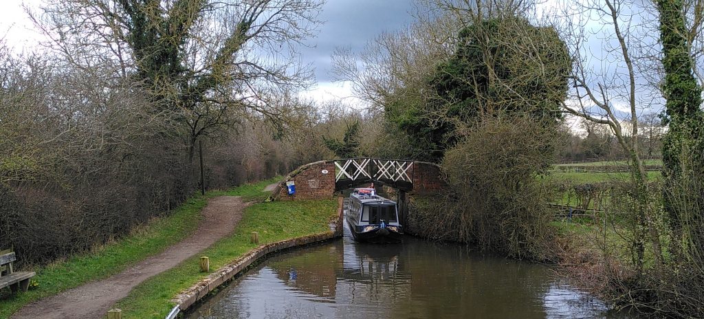 A narrowboat coming through a narrow bridge.  The bridge has brick sides with a black and white criss-cross pattern on the span.  The canal passes through fields and trees, some of which are still bare.