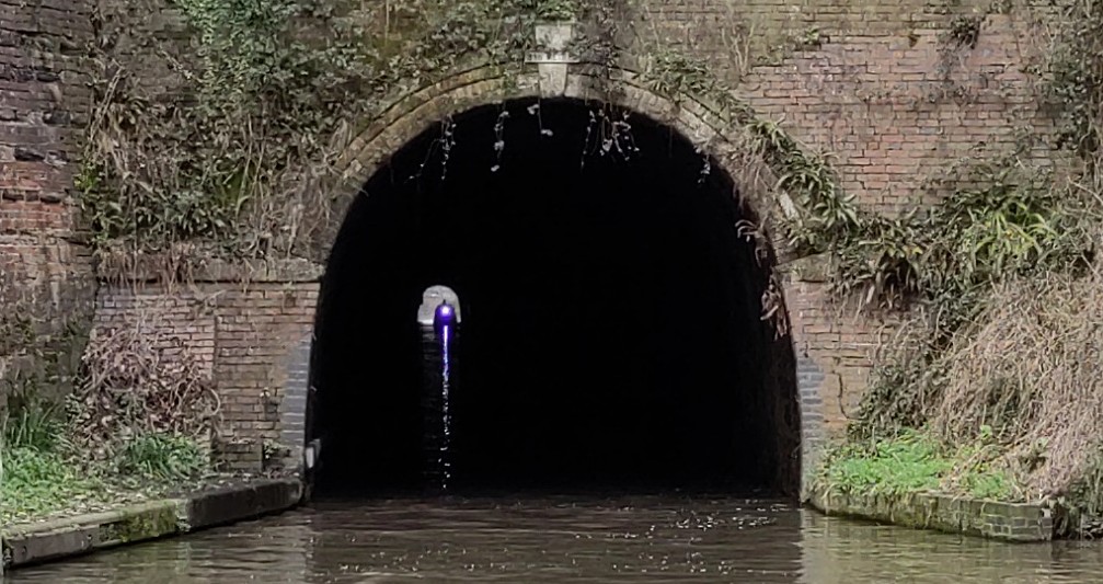 A canal tunnel portal.  The outline of a boat entering the far end is visible. Its light is reflected on the water for the whole length of the tunnel.