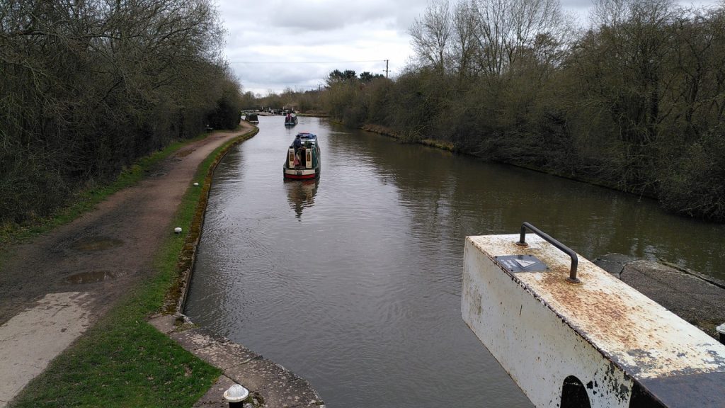 Two narrowboats receding along the canal. In the foreground is the end of an open lock gate arm.