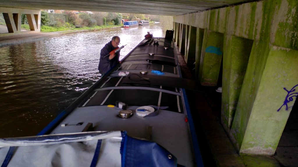 Narrowboat moored under a large concrete bridge. On the water side gunwale a man holds some equipment. Various other work equipment is also on the roof.