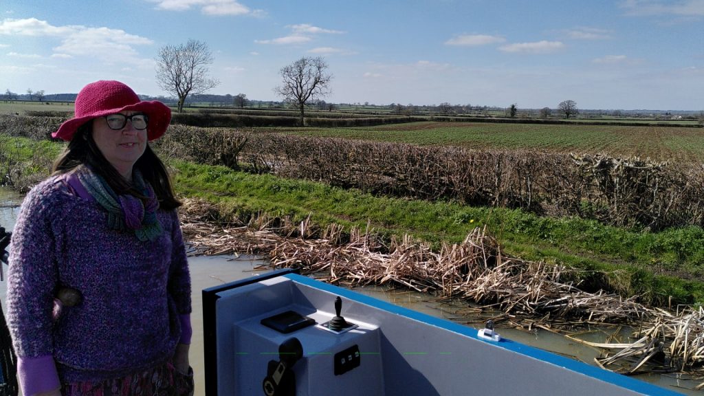 A narrowboater in a large red sun hat drives on a canal in a flat landscape.