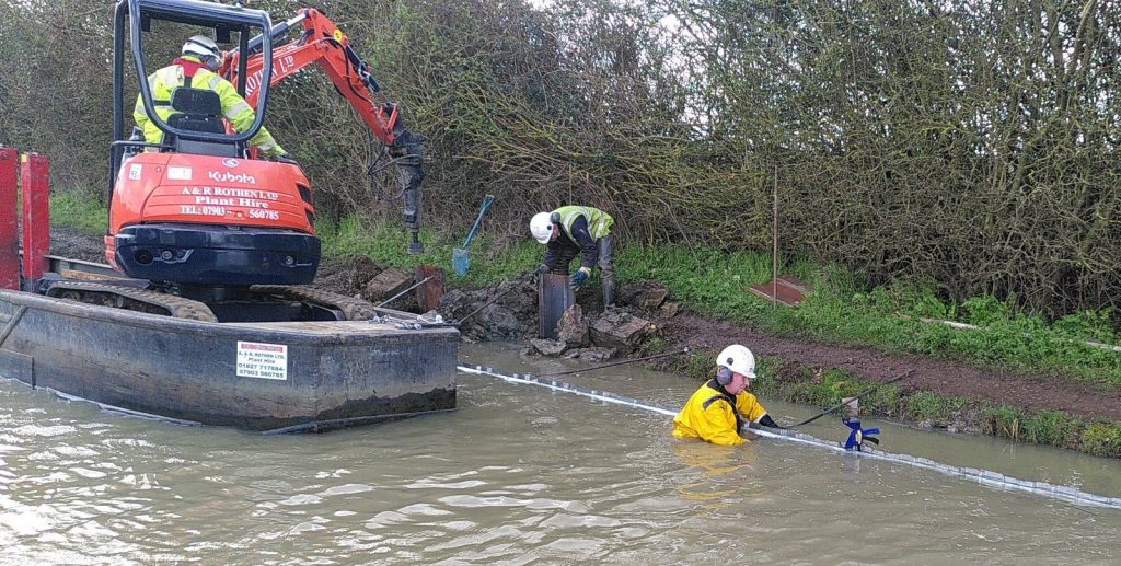 Three workers improving the canal towpath.  One is operating a digging machine sitting in a barge, another is standing on the towpath. The third is up to his shoulders in the muddy water of the canal.