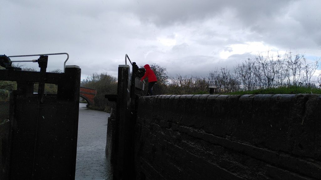 Viewed from inside a lock a figure in a red coat is closing the second gate.  The canal water visible through the gates is stippled with raindrops.