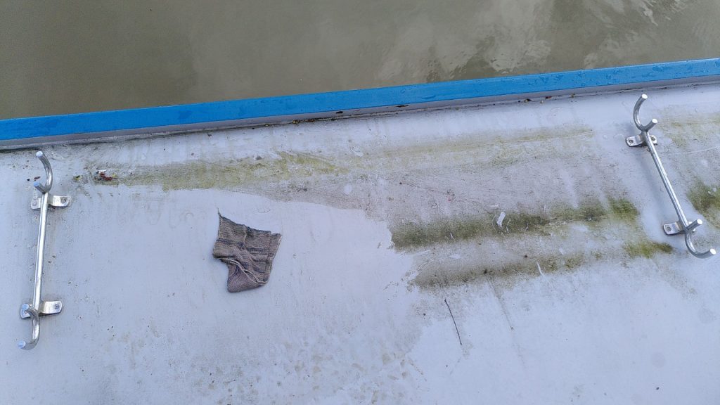 A narrowboat roof in the middle of being cleaned.  There is a build up of green weed in lines on the grey paint.  A cloth is dumped in the middle of the cleaned section.