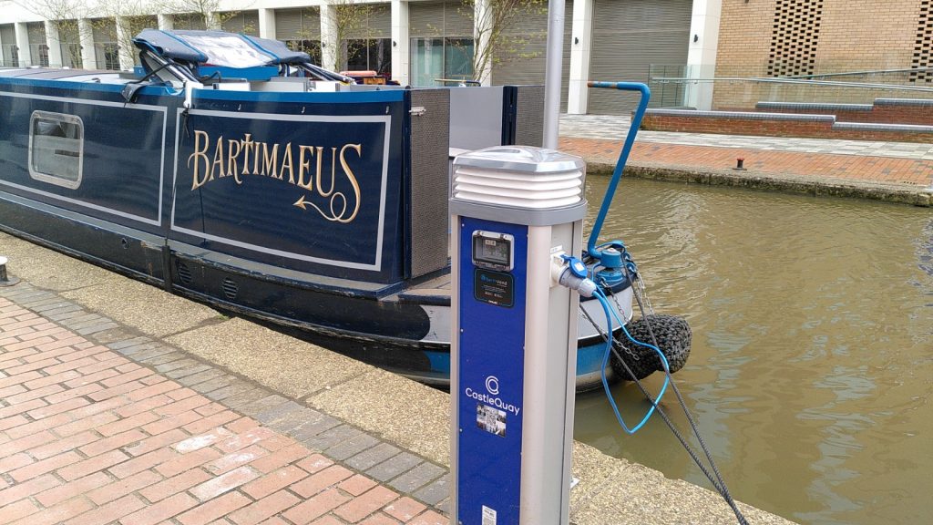 A charging point for narrowboats on a brick and concrete wharf. A cable leads to a moored narrowboat in the canal.