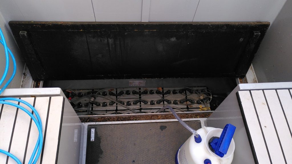 Stern deck of narrowboat with a panel lifted showing dozens of battery terminals across the width of the boat.  A small hose runs in to the compartment from a large plastic bottle.