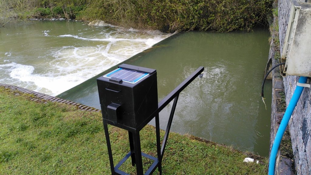 A metallic cube on stilts with solar panels on top sits beside a river. An arm protrudes over the river with a sensor on the end. Just downstream, the river flows over a weir producing foam downstream.