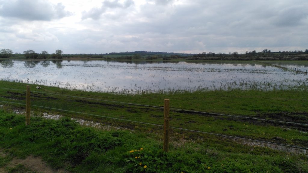 A view over a field with low hills beyond.  The main part of the field is flooded making it look like a small lake.