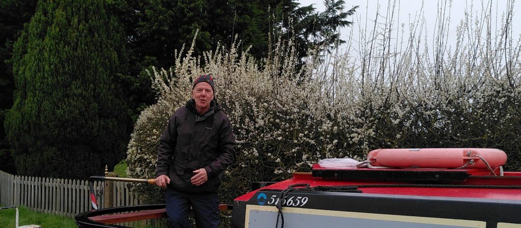 A man dressed mainly in dark clothes sits at the controls of a narrowboat. Behind is a spray of hawthorn blossom.