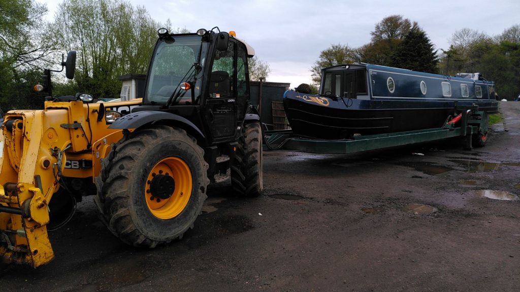 A yellow tractor behind which is a skeleton trailer with large balloon tyres. A narrowboat rests on the trailer.