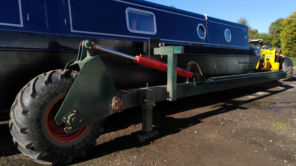 A large balloon tyre and hydraulic gear of a large trolley partially obscure a narrowboat on the trolley.  A face can be seen at a window of the boat.