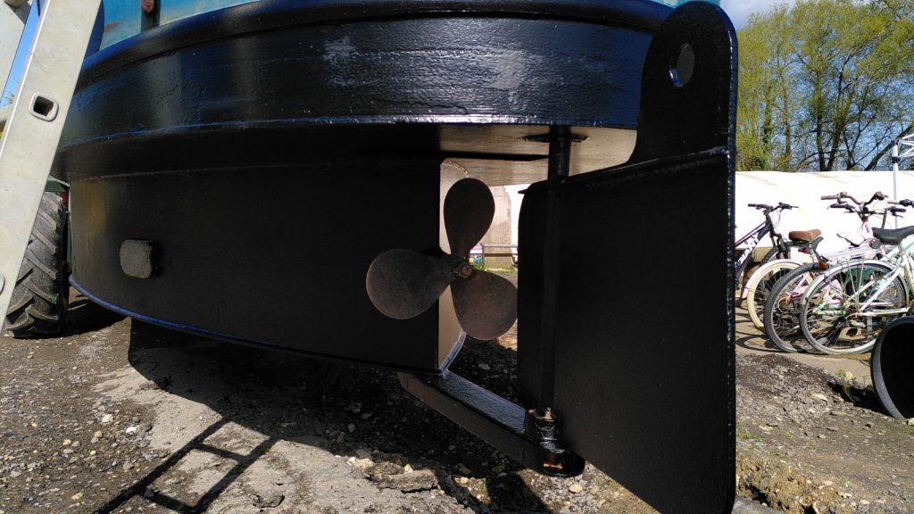 View under the stern of a narrowboat from behind. Beyond the rudder, the propellor sits.  The vertical hull sides meet at a point beyond the propellor.  Everything is freshly painted black.