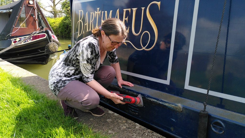 The side of a narrowboat with theartimaeus in large letters. Kneeling beside it, Clare is sanding below the gunwale.