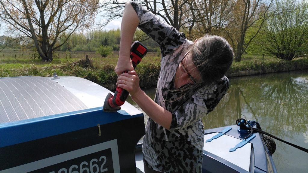 A woman standing in the fore-deck of a narrowboat sanding the top of the rail at the front of the cabin.