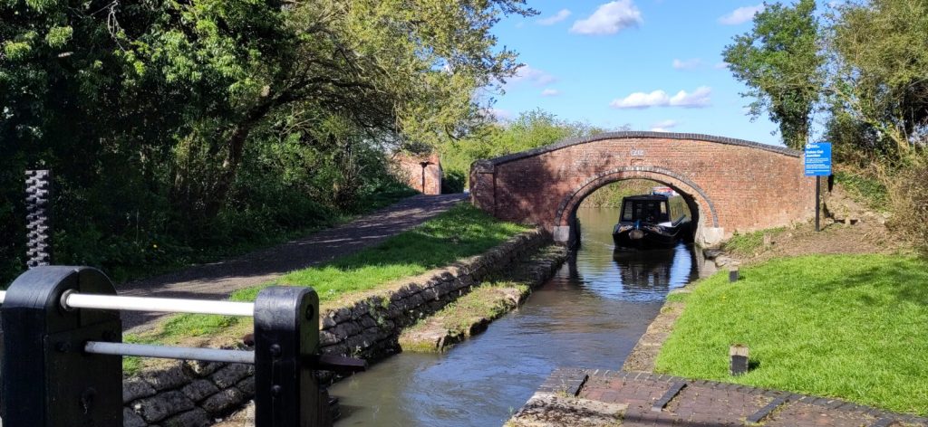 A narrow stone lined canal disappears under a brick arch bridge.  A narrowboat is coming towards us under the bridge.  It will have to stop at the lock gate, just visible in the foreground. 
