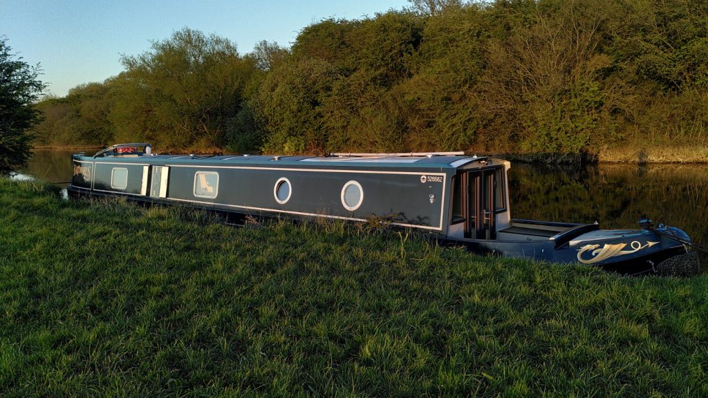 A narrowboat moored at the side of a field.  The gunwale of the narrowboat is at the same level as the field. The opposite bank is tree-lined.