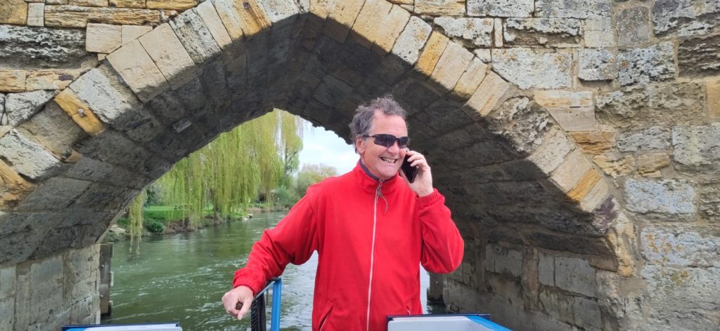 A man is steering a narrowboat while talking on a mobile phone. The boat has just come through a pointed arch on a stone bridge.
