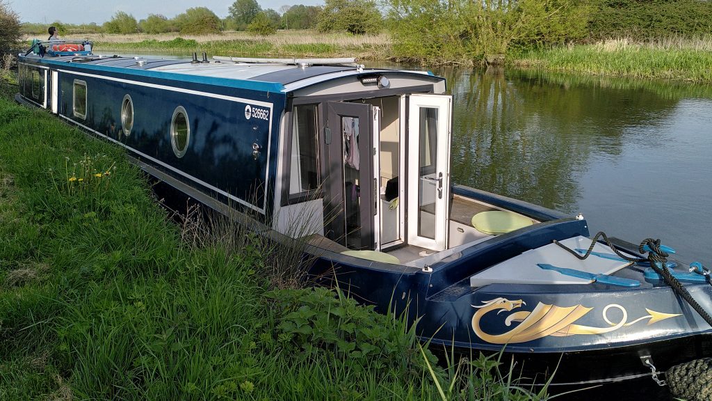 A narrowboat is moored against the bank of a river. The near bank is at the height of the gunwale. The opposite bank is green and slightly lower.