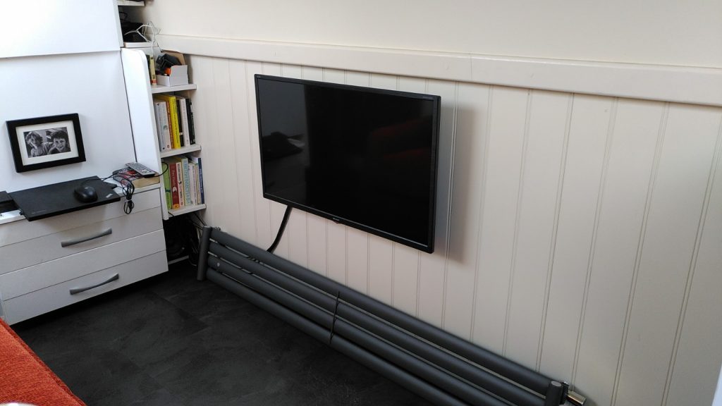 The cabin of a narrowboat with a TV on a side wall. A sofa is just visible facing it.  There is a wide short radiator on the floor under the TV.