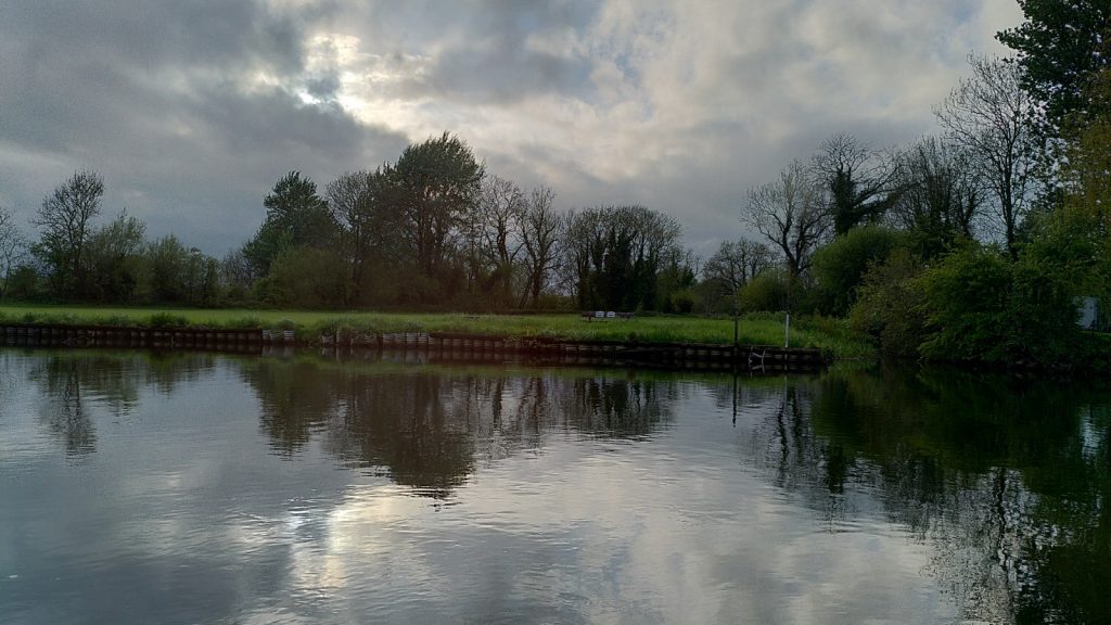 A view across the river.  The field opposite is flat with trees behind.  The sky has heavy clouds with some  brightness breaking through.  The entire scene is reflected in the river.