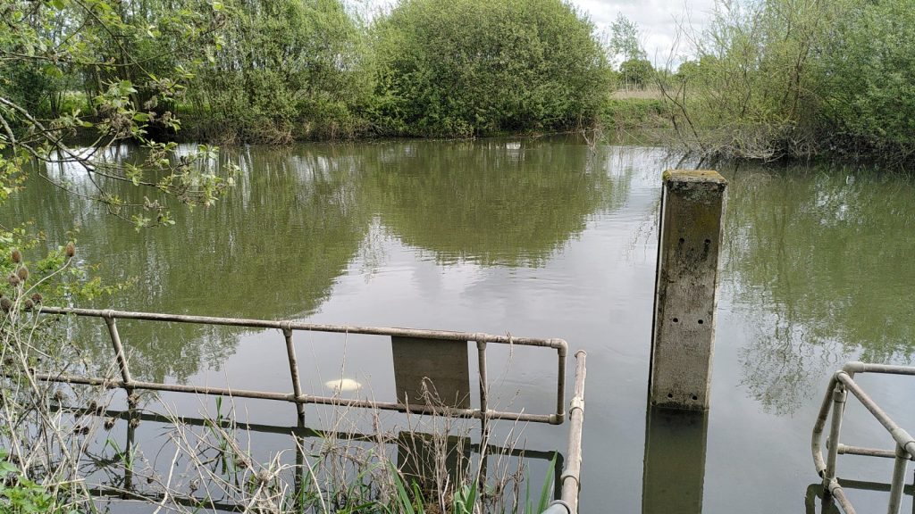 View across a canal.  On the near side is the lock landing.  It is completely submerged.  A submerged bollard intended for tying up can just be seen.