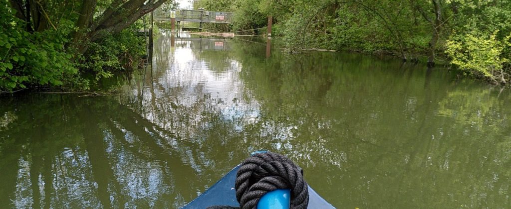 View over the moored nose of a narrowboat. A short tree lined section of canal stops is blocked by a weir with a gantry above on which a large sign says DANGER.